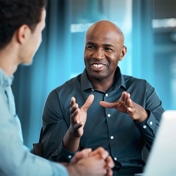 Two men sit at a table and have a conversation. One man gestures with his hands while speaking; the other listens. A laptop is on the table beside them.