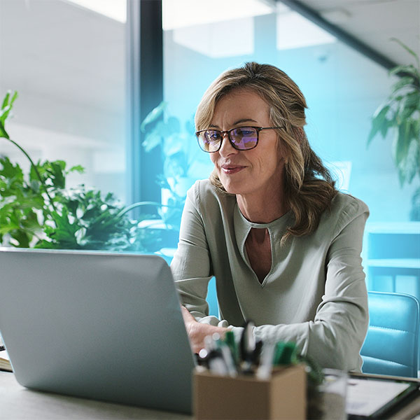 A woman wearing glasses sits at a desk working on a laptop in a modern office with plants in the background.
