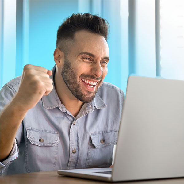 A man smiles and raises his fist in excitement while looking at a laptop screen in a bright room.